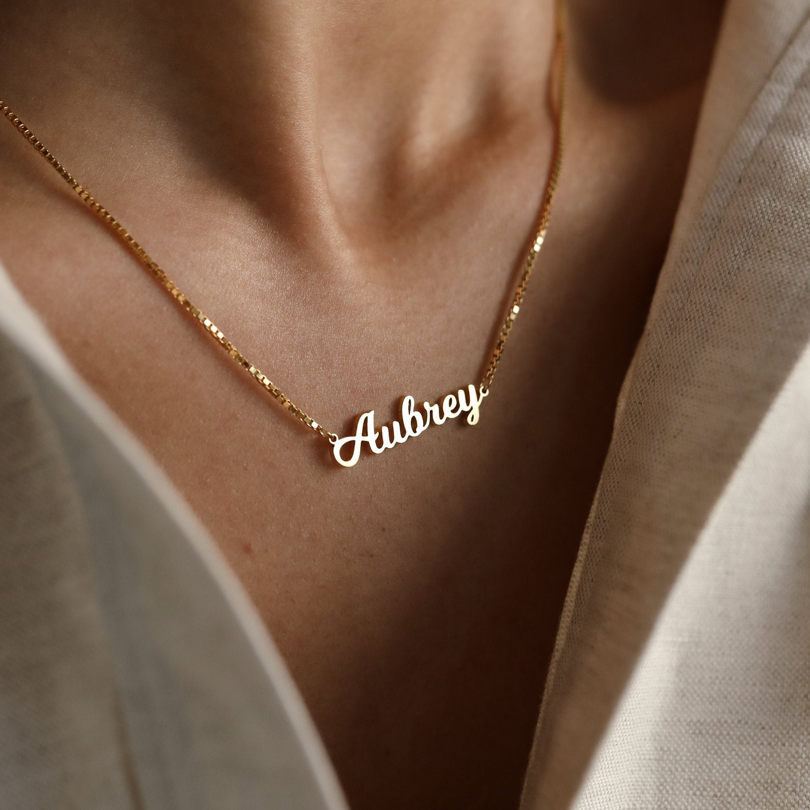 A Mellow Name Necklace in Box Chain with "Aubrey" rests on a person's neck, its elegant chain visible above a cream-colored shirt.
