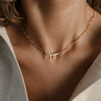 The Summer Name Necklace in Figaro Chain features a Maya nameplate in cursive font, displayed on a woman's neck with a delicate gold Figaro chain, shown in close-up.