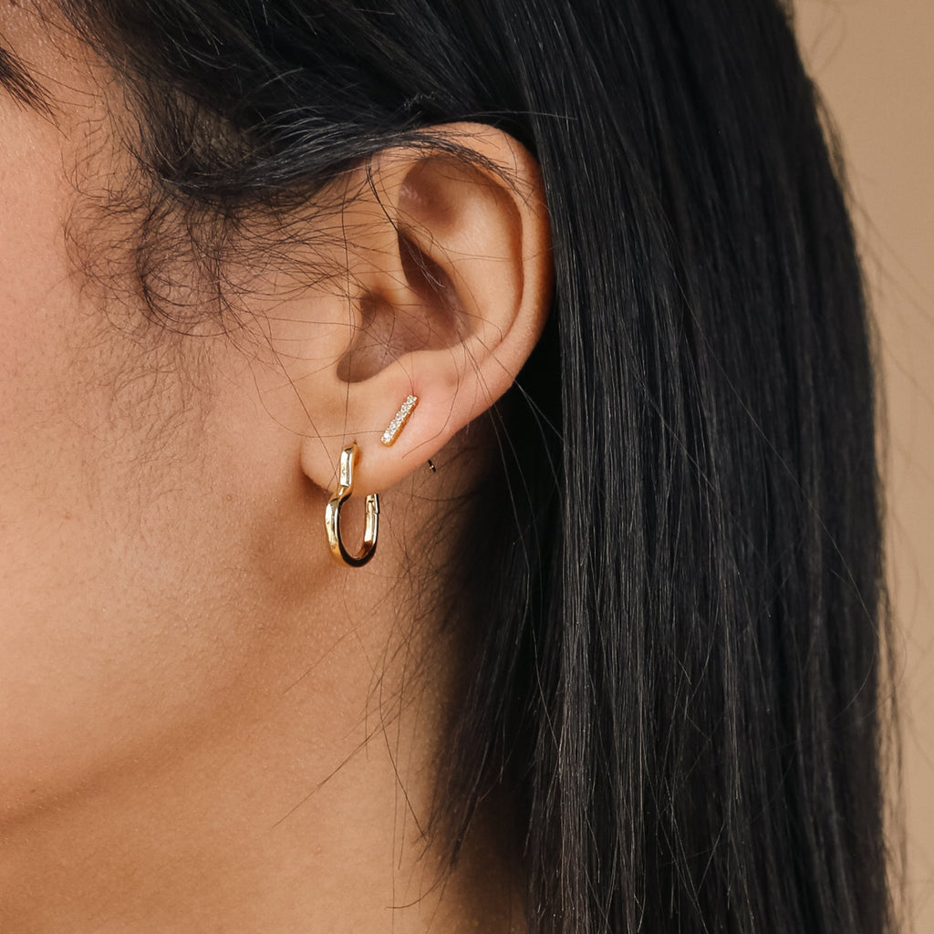 Close-up of an ear wearing two small gold hoop earrings, featuring the Heart Hoops design, along with a small gold bar stud on the earlobe.