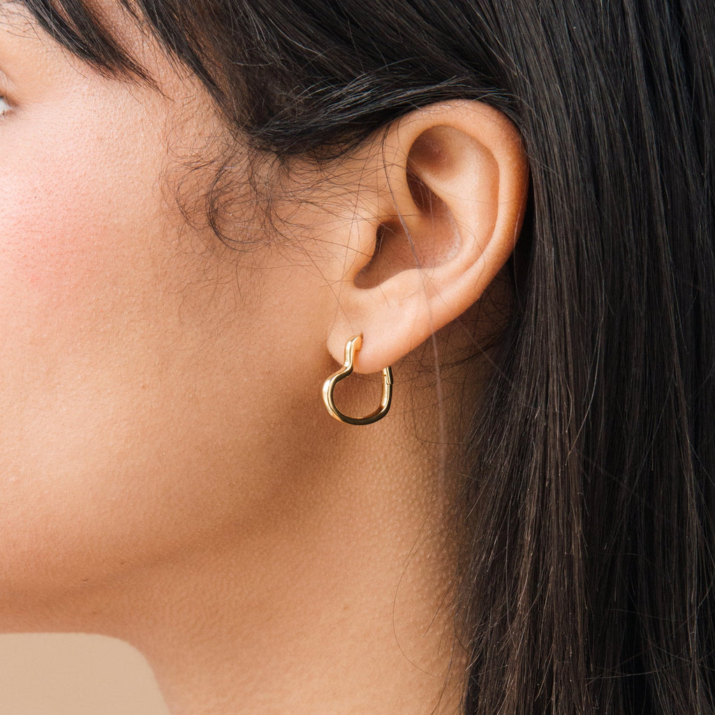 Close-up of a woman's ear wearing the Heart Hoops, small gold heart-shaped earrings; her straight dark hair is partially visible.