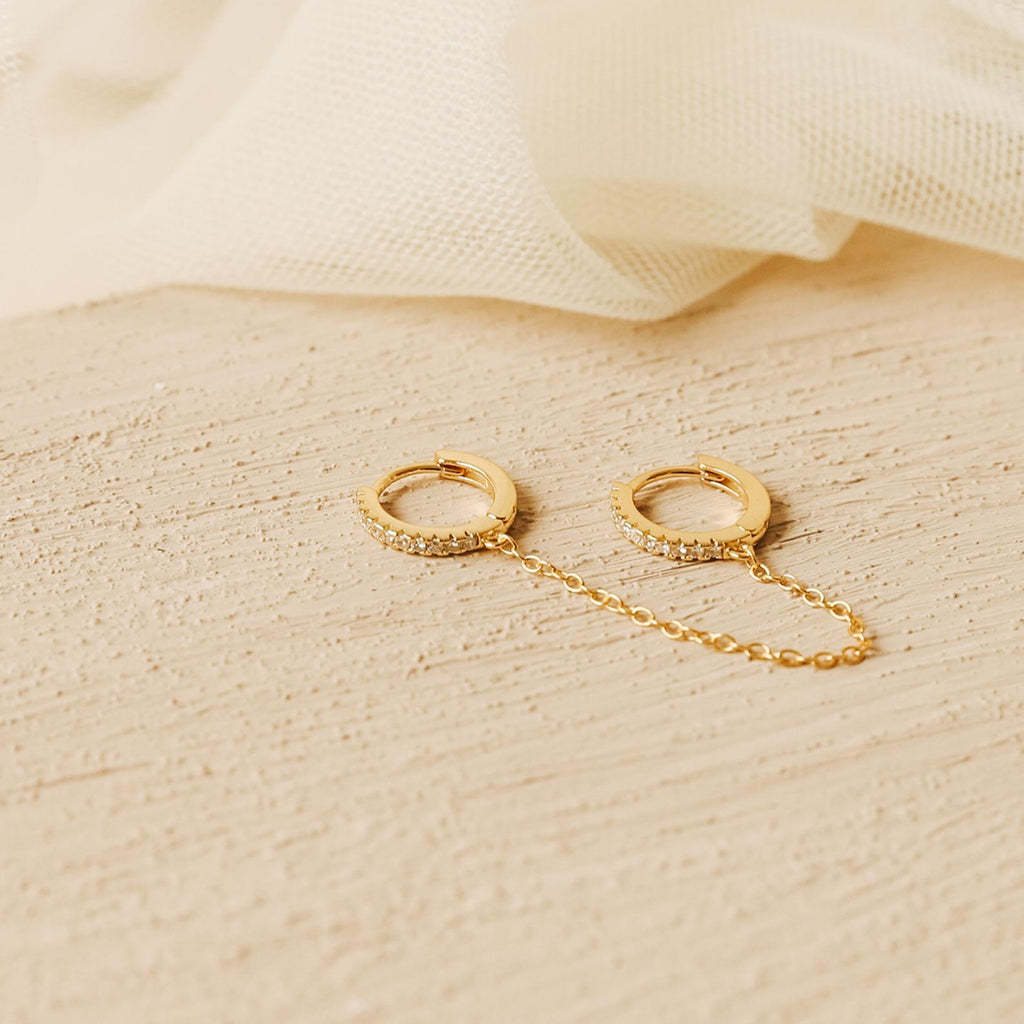 Two Pave Duo Chain Earrings, ideal for multiple ear piercings, are linked by a delicate chain and displayed on a textured beige surface with soft fabric in the background.