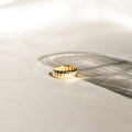 A gold Audrey Ring with a grooved pattern rests on a sunlit surface, casting a gentle shadow—an essential piece ideal for everyday wear.