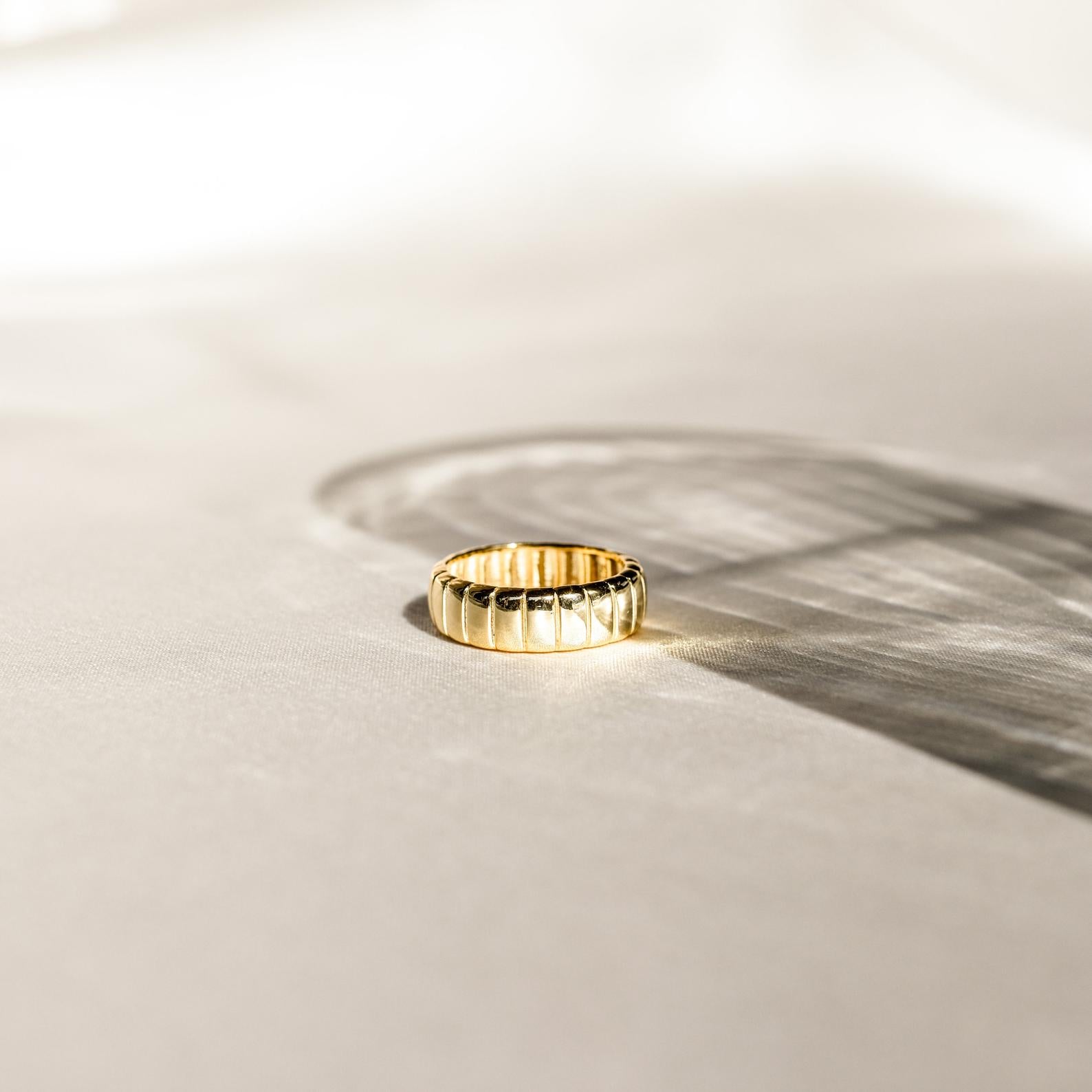 A gold Audrey Ring with a grooved pattern rests on a sunlit surface, casting a gentle shadow—an essential piece ideal for everyday wear.