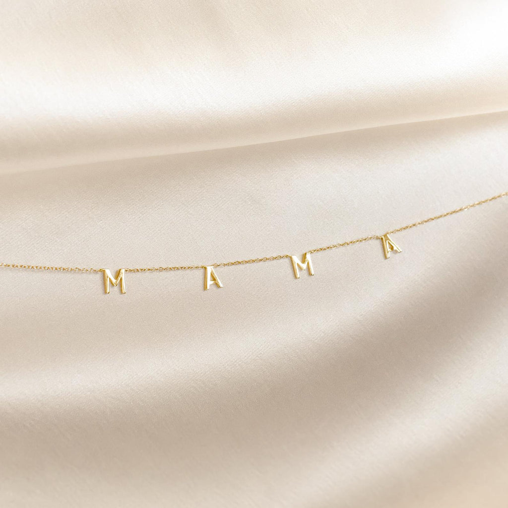 The Mama Letter Necklace, with charms spelling MAMA, is displayed on soft cream-colored fabric—a perfect gift to celebrate motherhood.