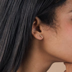 A closeup of a woman's ear with straight black hair showcases minimal jewelry—a small, horizontal gold Pave Bar Studs earring.