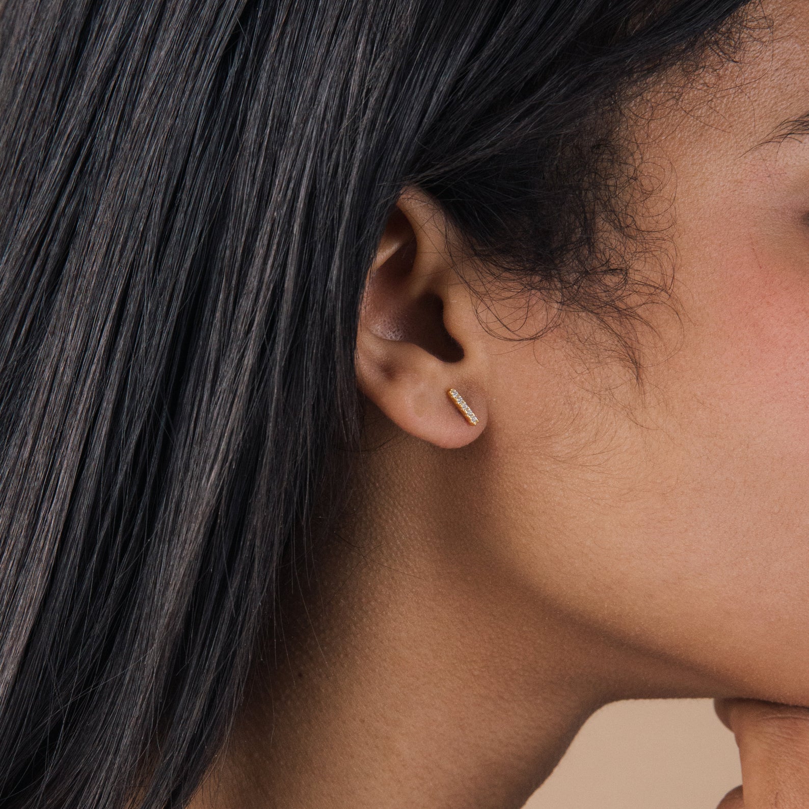 A closeup of a woman's ear with straight black hair showcases minimal jewelry—a small, horizontal gold Pave Bar Studs earring.
