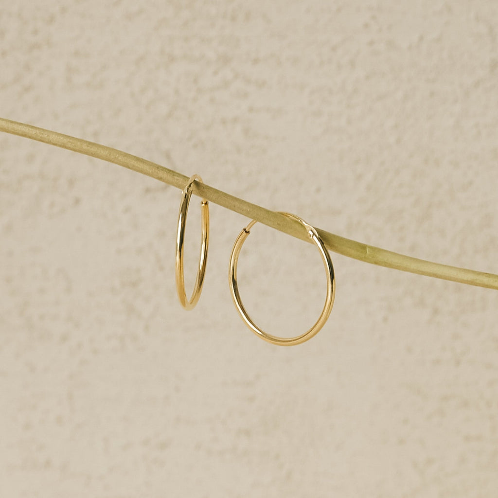 The Endless Thin Hoops, displayed on a slender pale branch with a textured beige background, are timeless gold earrings crafted to be the perfect everyday accessory.