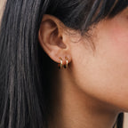 Close-up of a woman wearing the Twin Hoops—small gold earrings that create a subtle double earring illusion—partially hidden by her straight black hair.