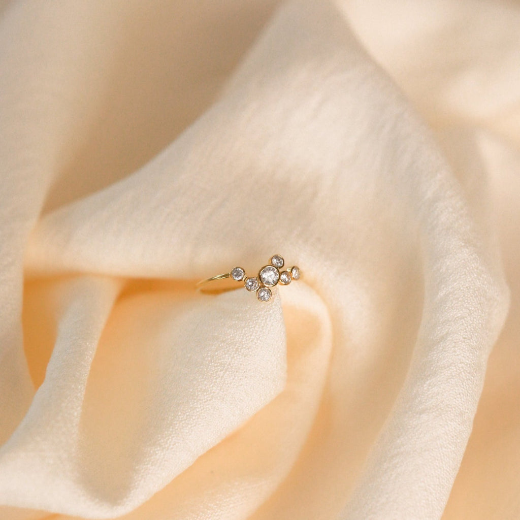 The Cluster Birthstone Ring, featuring a classic gold design with multiple small diamonds, rests on soft cream-colored fabric.