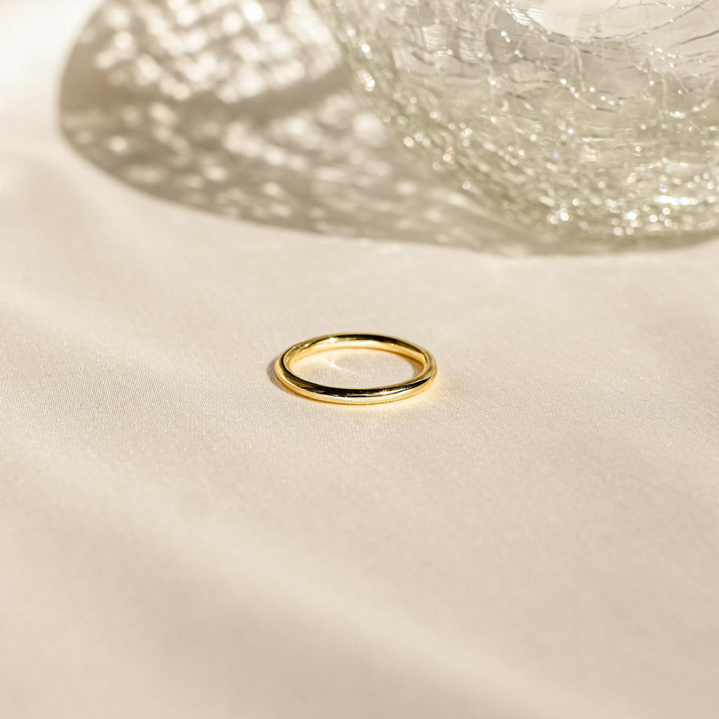 The Minimal Stacking Ring, a delicate gold piece, rests on cream fabric with textured glass casting shadows—perfect for stacking and everyday elegance.