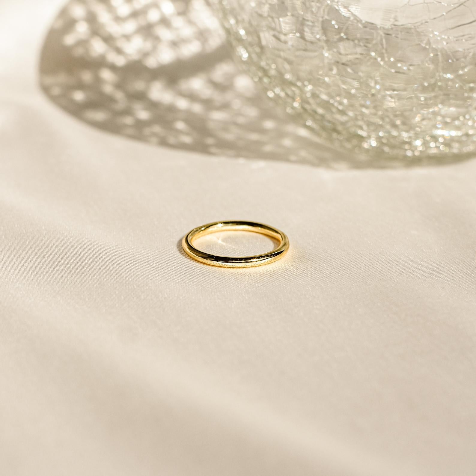 The Minimal Stacking Ring, a delicate gold piece, rests on cream fabric with textured glass casting shadows—perfect for stacking and everyday elegance.
