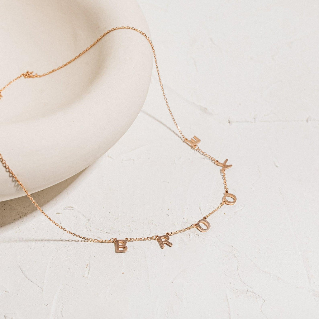 The Alice Letter Name Necklace in Rose Gold features dangling letter charms spelling BROKE on a textured surface near a round dish—a playful, personalized jewelry piece ideal for a unique Christmas gift.