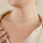 Minimalist gold necklace with a slim horizontal bar pendant encrusted with tiny clear stones, worn at the collarbone and styled with a light satin blouse.