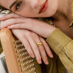 A woman in a green blouse rests her hands, adorned with the Bold Dome Ring, on a woven chair and gazes softly at the camera.