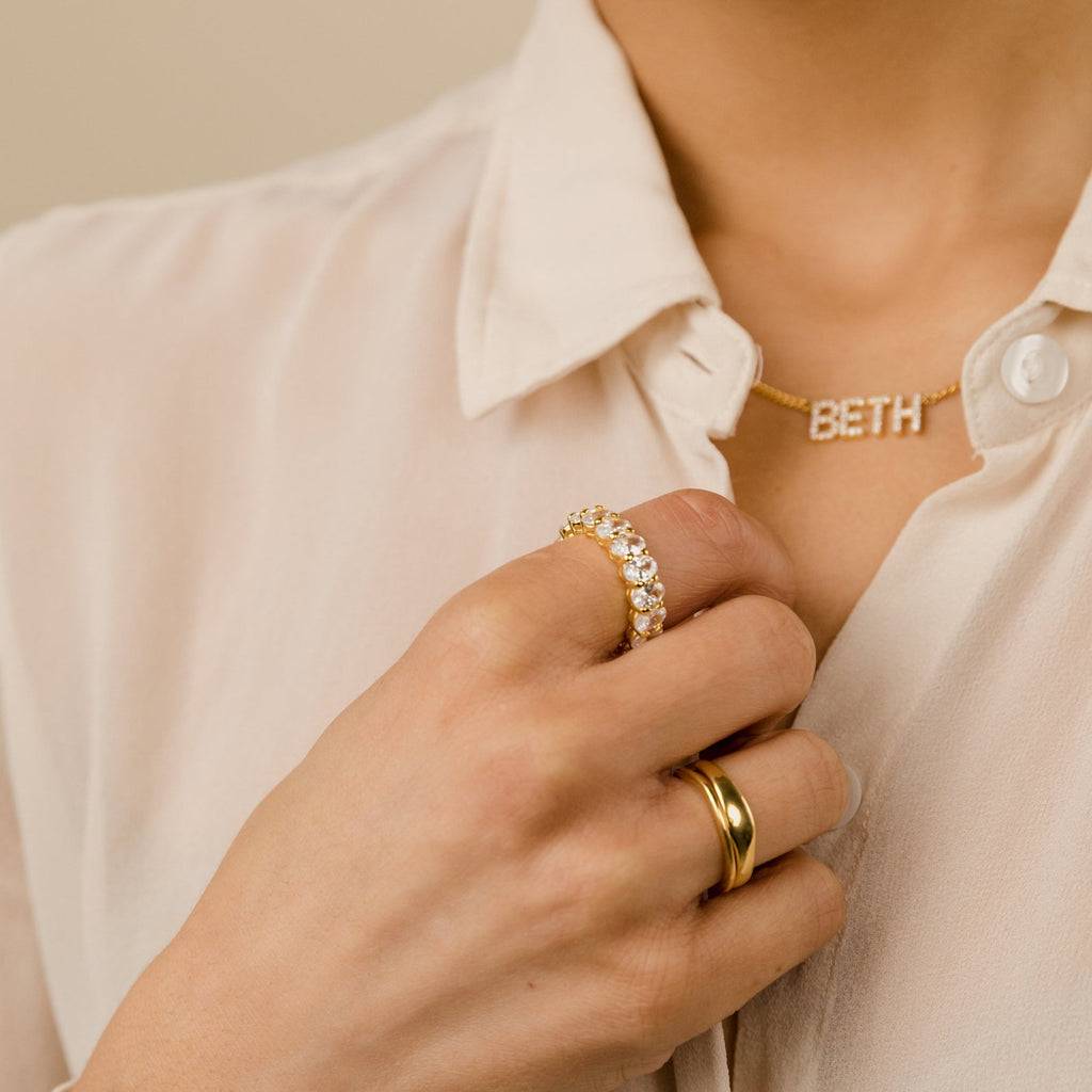 A person wearing elegant jewelry, including the Alannah Ring and a gold necklace with letter charms spelling BETH, over a cream blouse.