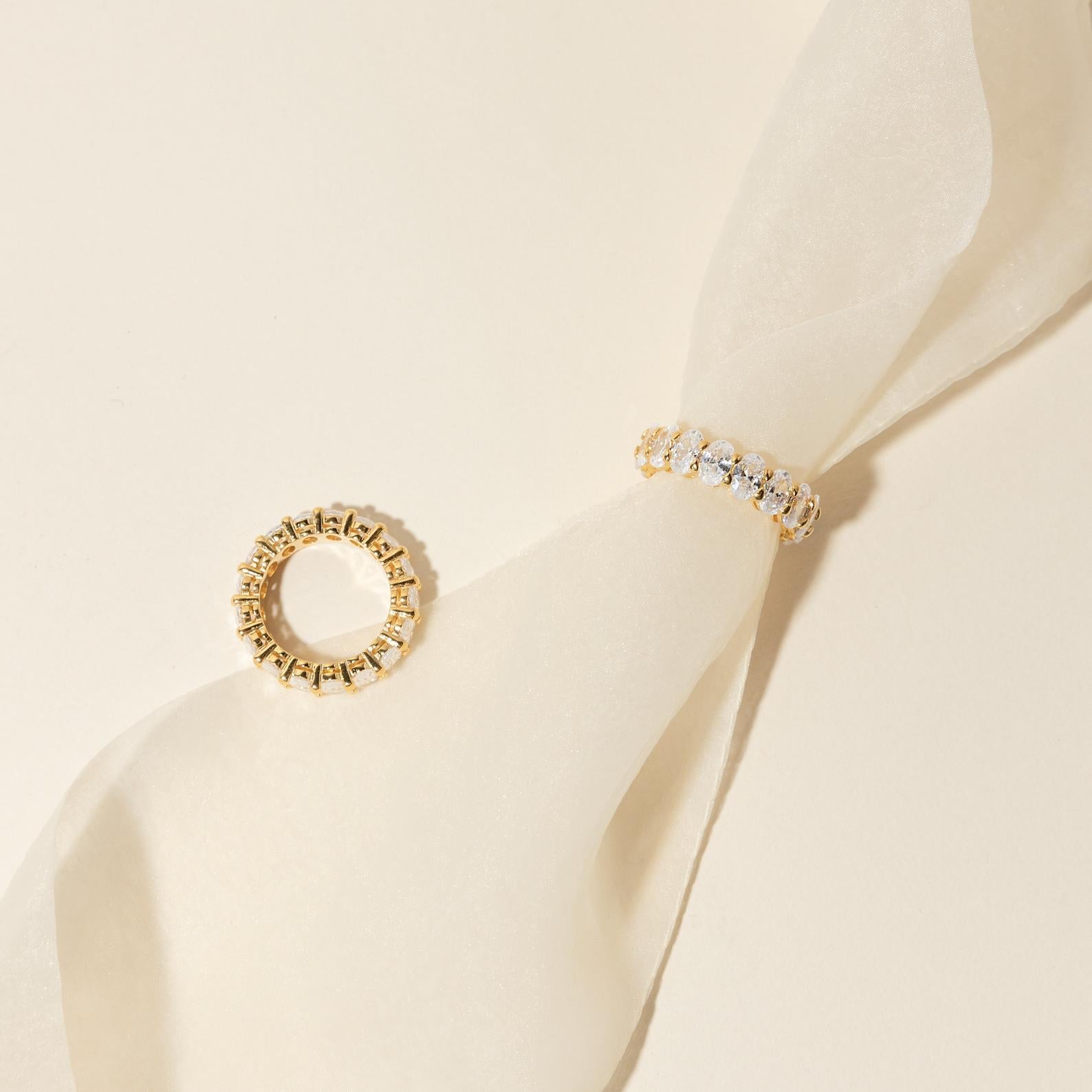 The Alannah Ring, a gold oval diamond ring paired with a shimmering infinity band, is displayed on a light, sheer fabric background.