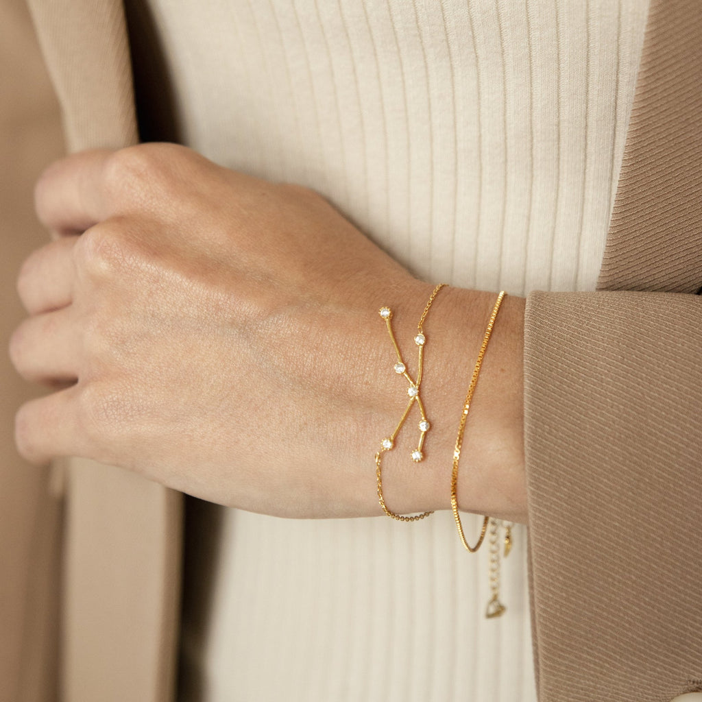 A hand wears the delicate gold Constellation Bracelet with small pearls, styled with two other pearl bracelets against a beige outfit and coat for a minimal look.