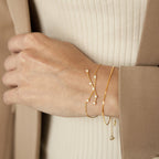 A hand wears the delicate gold Constellation Bracelet with small pearls, styled with two other pearl bracelets against a beige outfit and coat for a minimal look.