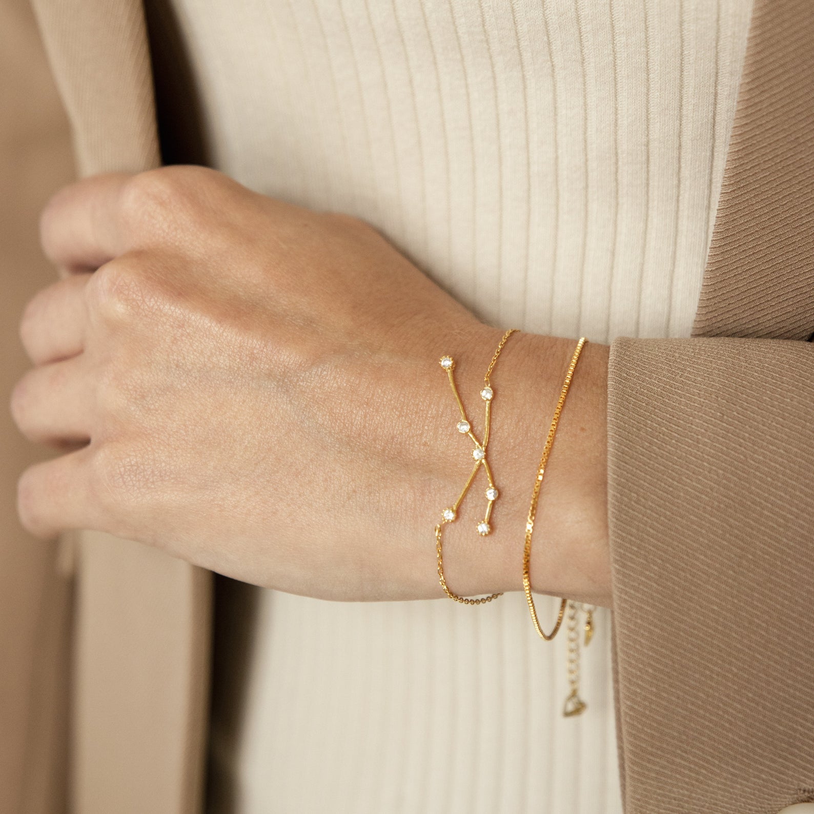 A hand wears the delicate gold Constellation Bracelet with small pearls, styled with two other pearl bracelets against a beige outfit and coat for a minimal look.