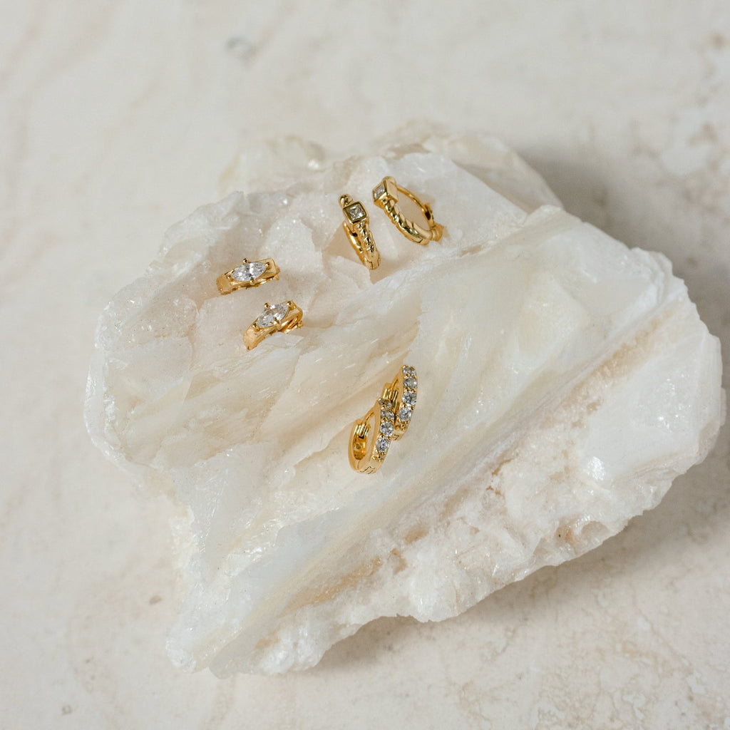 Five Aurora Huggies, featuring various designs and intricate diamond details, displayed on a large, textured white crystal stone.