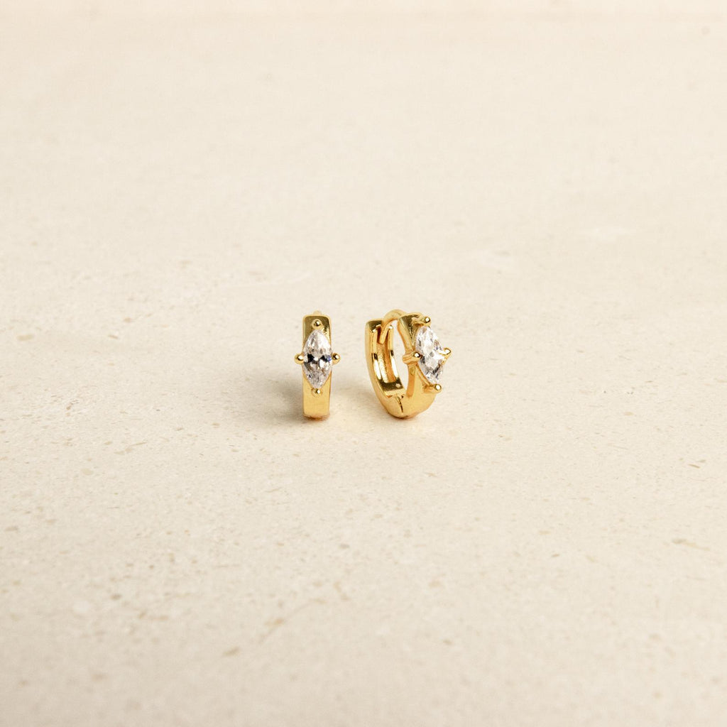 The Aurora Huggies are dainty small gold hoop earrings with marquise-shaped clear stones and subtle diamond accents, shown on a light beige surface.