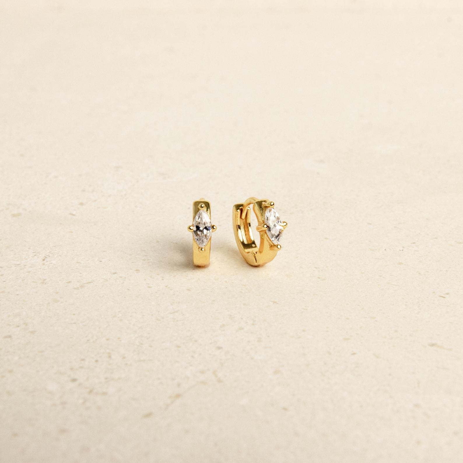 The Aurora Huggies are dainty small gold hoop earrings with marquise-shaped clear stones and subtle diamond accents, shown on a light beige surface.