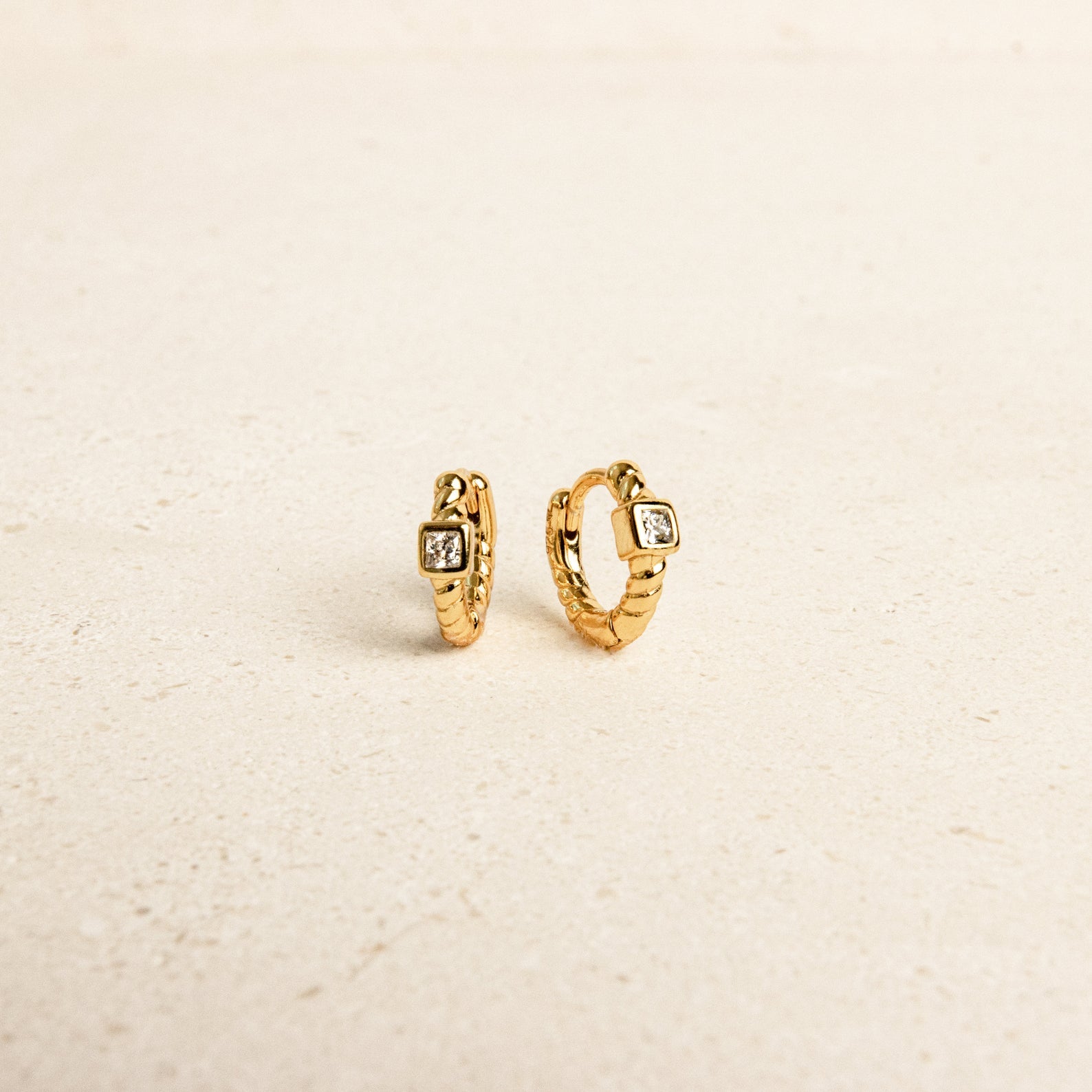 The Willow Huggies are small gold hoop earrings with a square gemstone, crafted from 925 Sterling Silver and displayed on a light beige surface.
