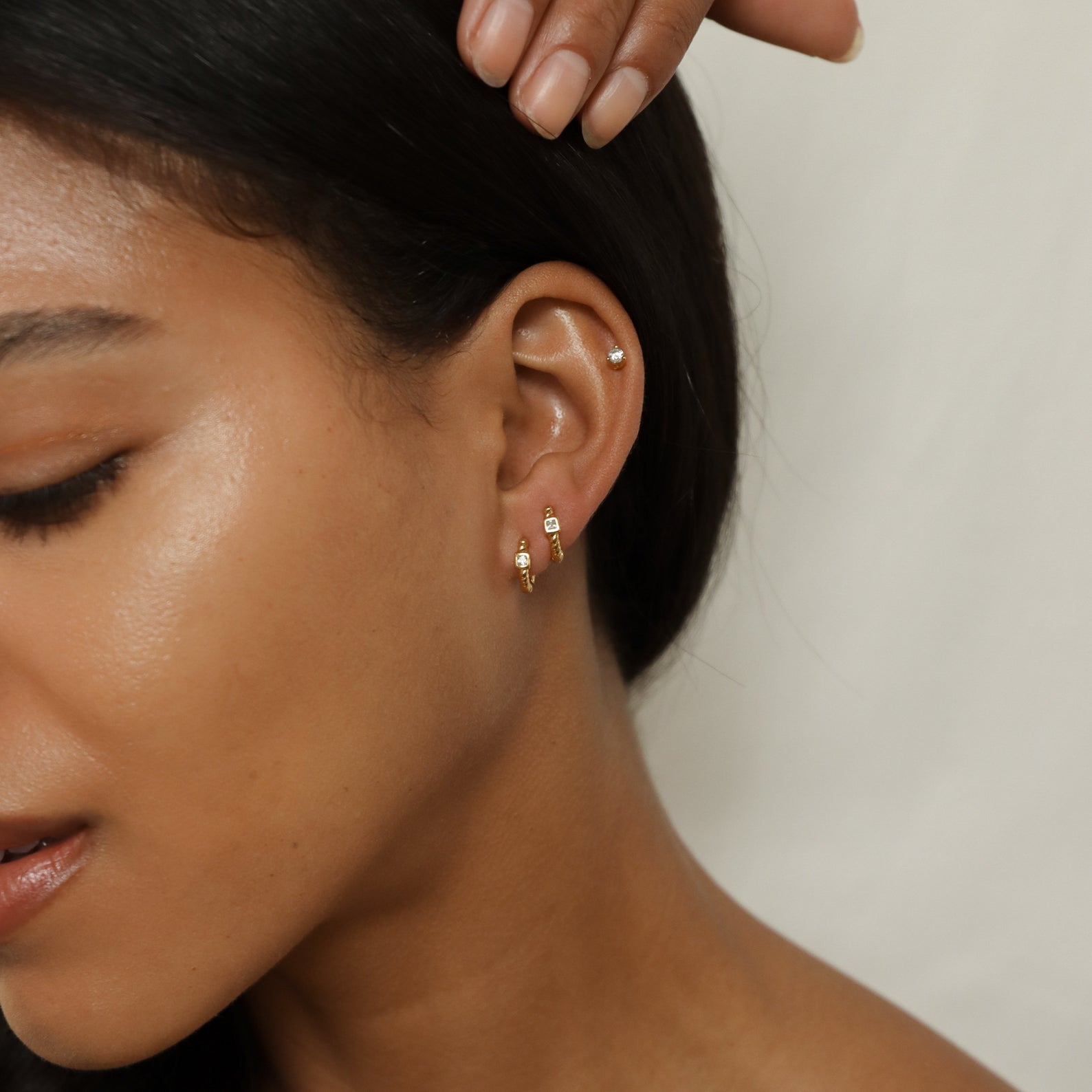 Close-up of a woman’s ear adorned with three gold hoop earrings and one small stud, her hand touching her hair. The Willow Huggies, crafted in 925 sterling silver, add a touch of elegance to her look.