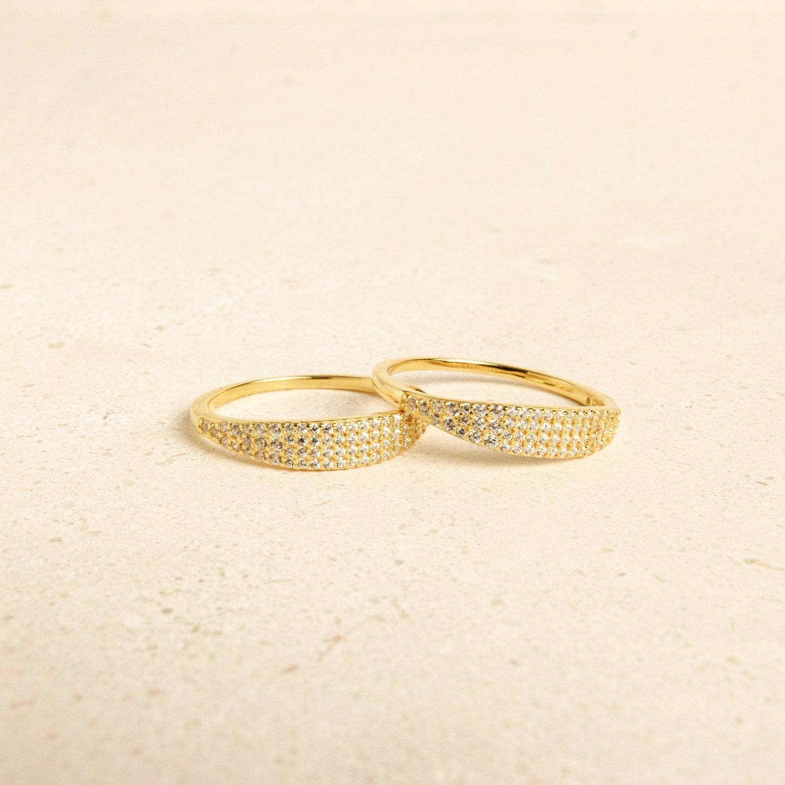 Two Gina Pave Signet Rings, featuring gold bands accented with small sparkling stones, are placed side by side on a light, textured surface.