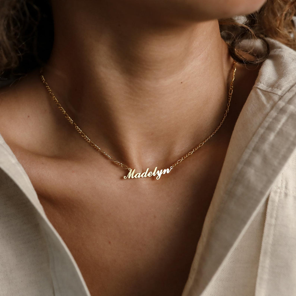A woman wears the Carrie Name Necklace in Figaro Chain, featuring the name Madelyn in cursive gold letters.