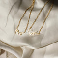 Two Carrie Name Necklaces in Figaro chains, customized with the names Maya and Sophia, rest gracefully on cream-colored satin fabric in an elegant minimal style.