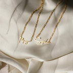 Two Carrie Name Necklaces in Figaro chains, customized with the names Maya and Sophia, rest gracefully on cream-colored satin fabric in an elegant minimal style.