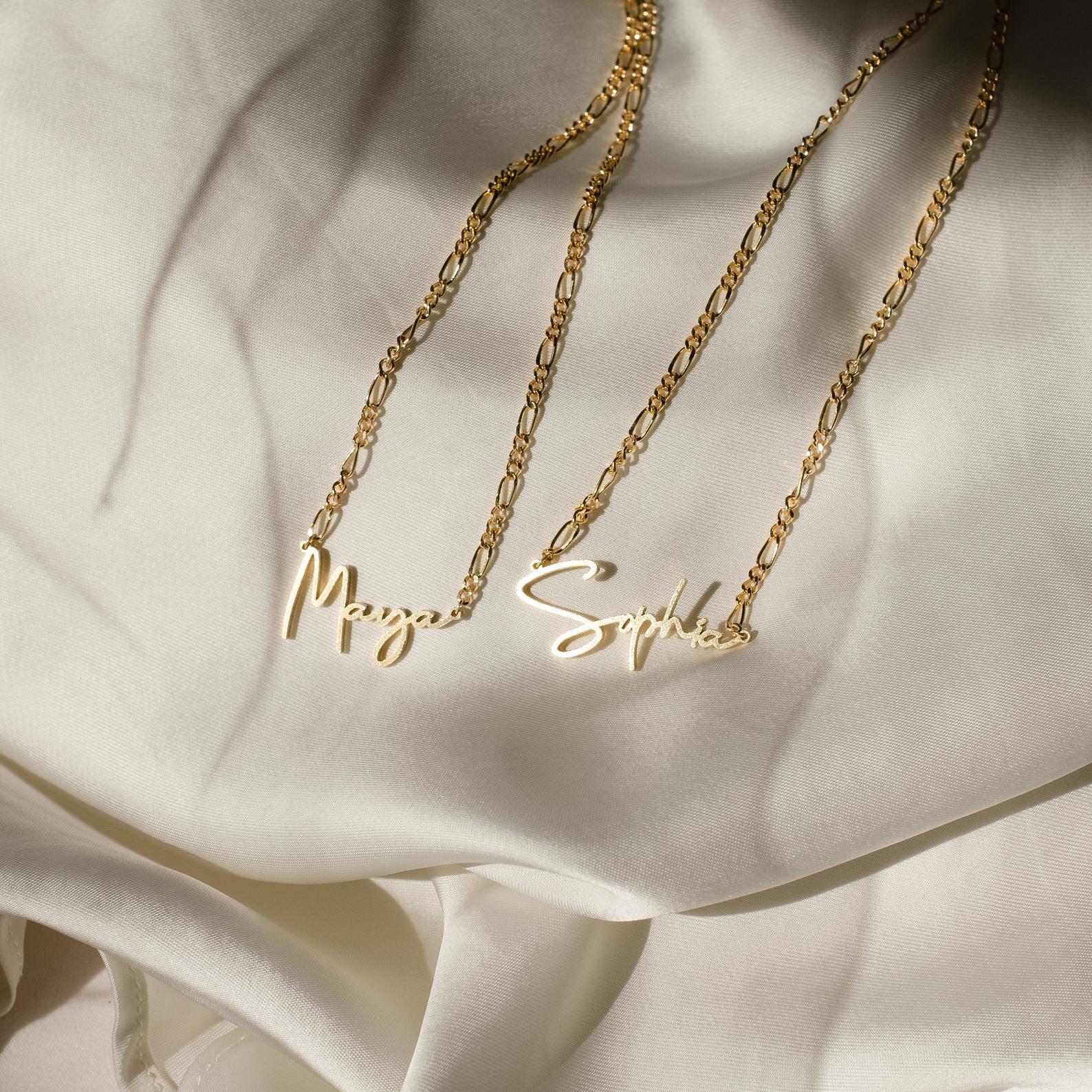 Two Carrie Name Necklaces in Figaro chains, customized with the names Maya and Sophia, rest gracefully on cream-colored satin fabric in an elegant minimal style.