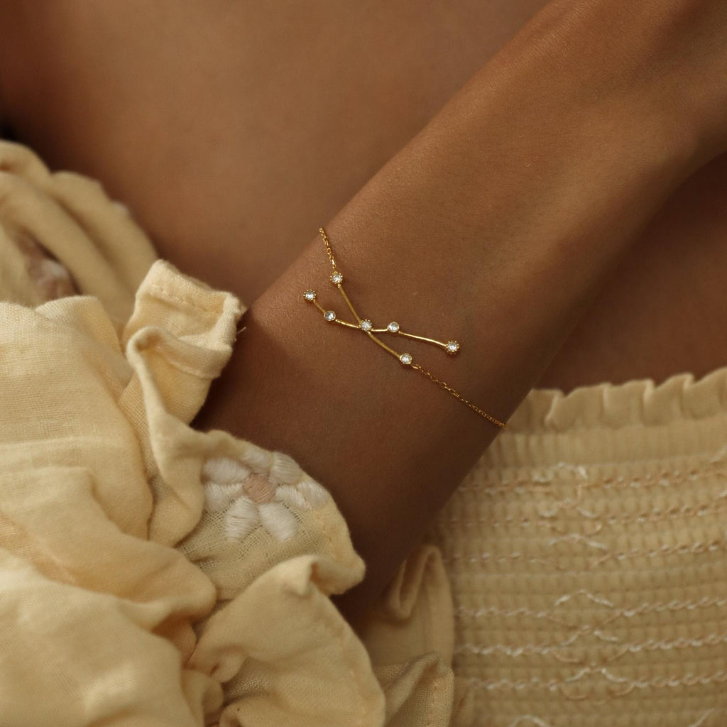The Constellation Bracelet features delicate branches and beads, shown on a woman's wrist with a textured cream dress—a minimal yet unique accessory for jewelry lovers.