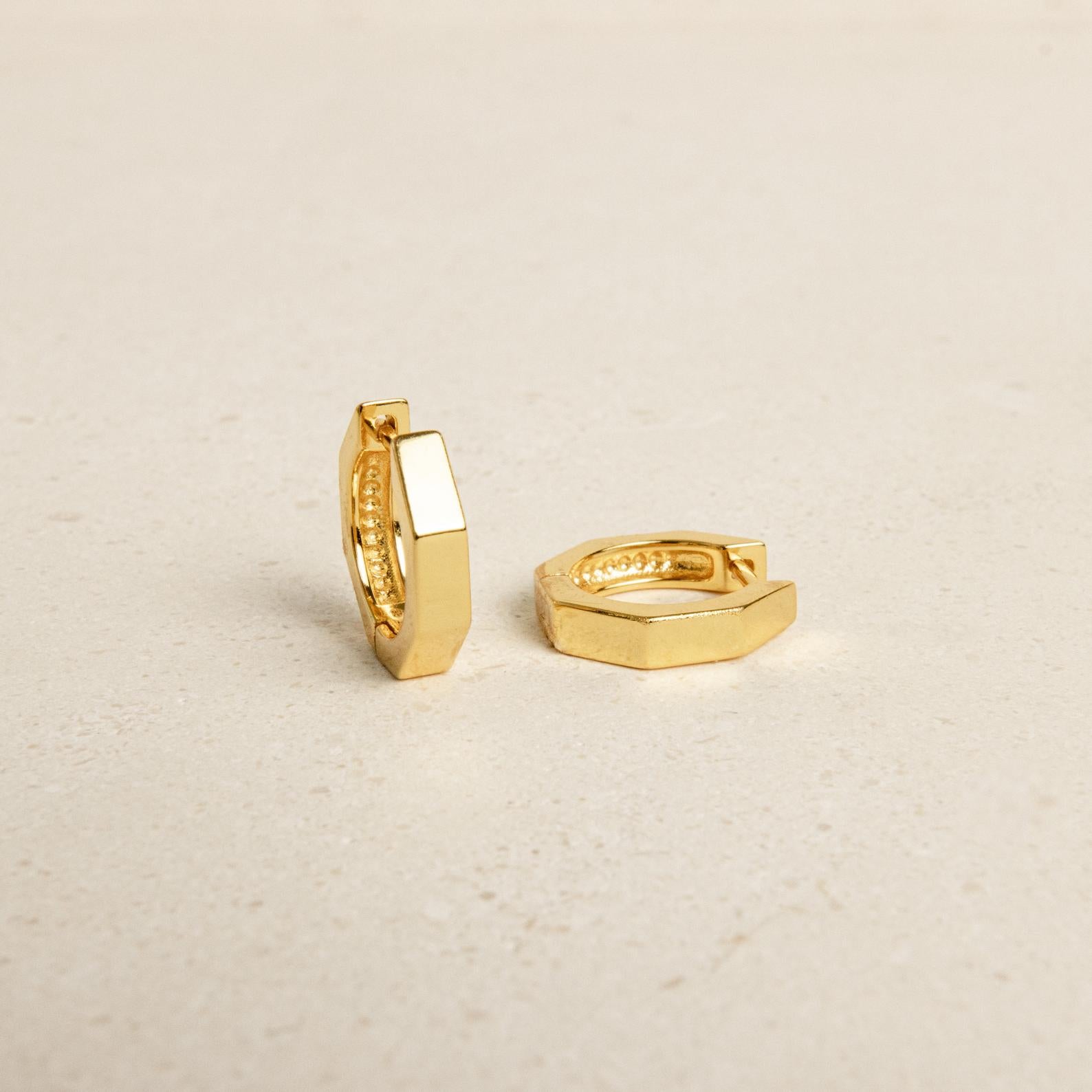 The Audrey Huggies are two small, gold-finished hexagon earrings resting on a light beige surface—an elegant everyday accessory perfect for any occasion.