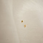The Lightning Bolt Studs, featuring a small gold design, rest on a sheer white fabric background—perfect for adding an edgy jewelry accent to your look.