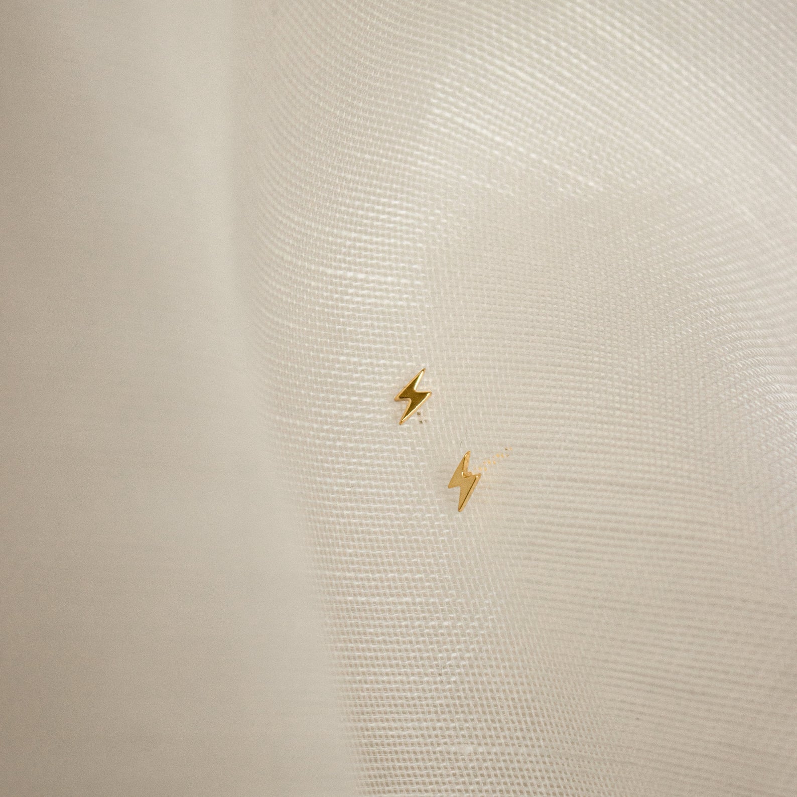 The Lightning Bolt Studs, featuring a small gold design, rest on a sheer white fabric background—perfect for adding an edgy jewelry accent to your look.