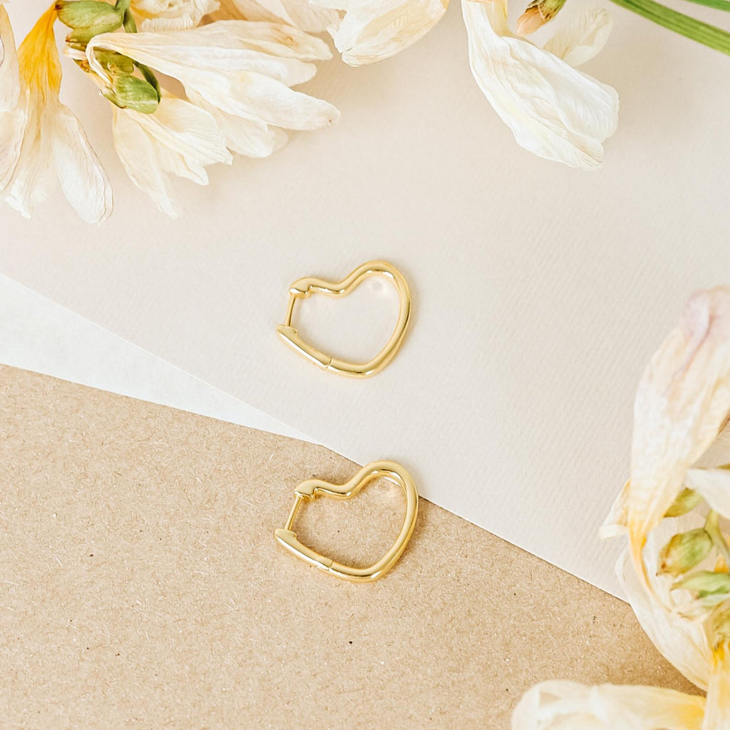 The Heart Hoops are gold heart-shaped earrings displayed on paper with cream-colored flowers, featuring a delicate open-heart design.