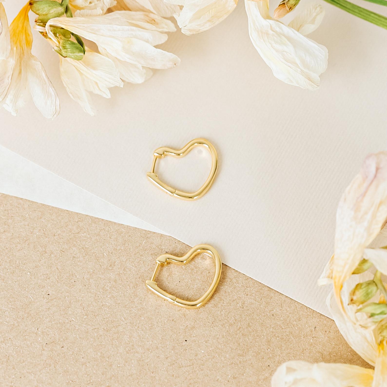 The Heart Hoops are gold heart-shaped earrings displayed on paper with cream-colored flowers, featuring a delicate open-heart design.