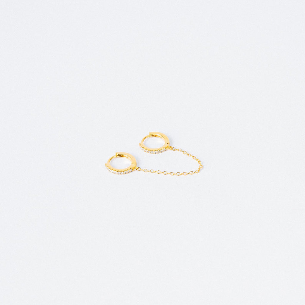 The Pave Duo Chain Earrings feature two small gold huggies linked by a delicate chain, with one hoop embellished with clear stones—perfect for styling with multiple ear piercings.