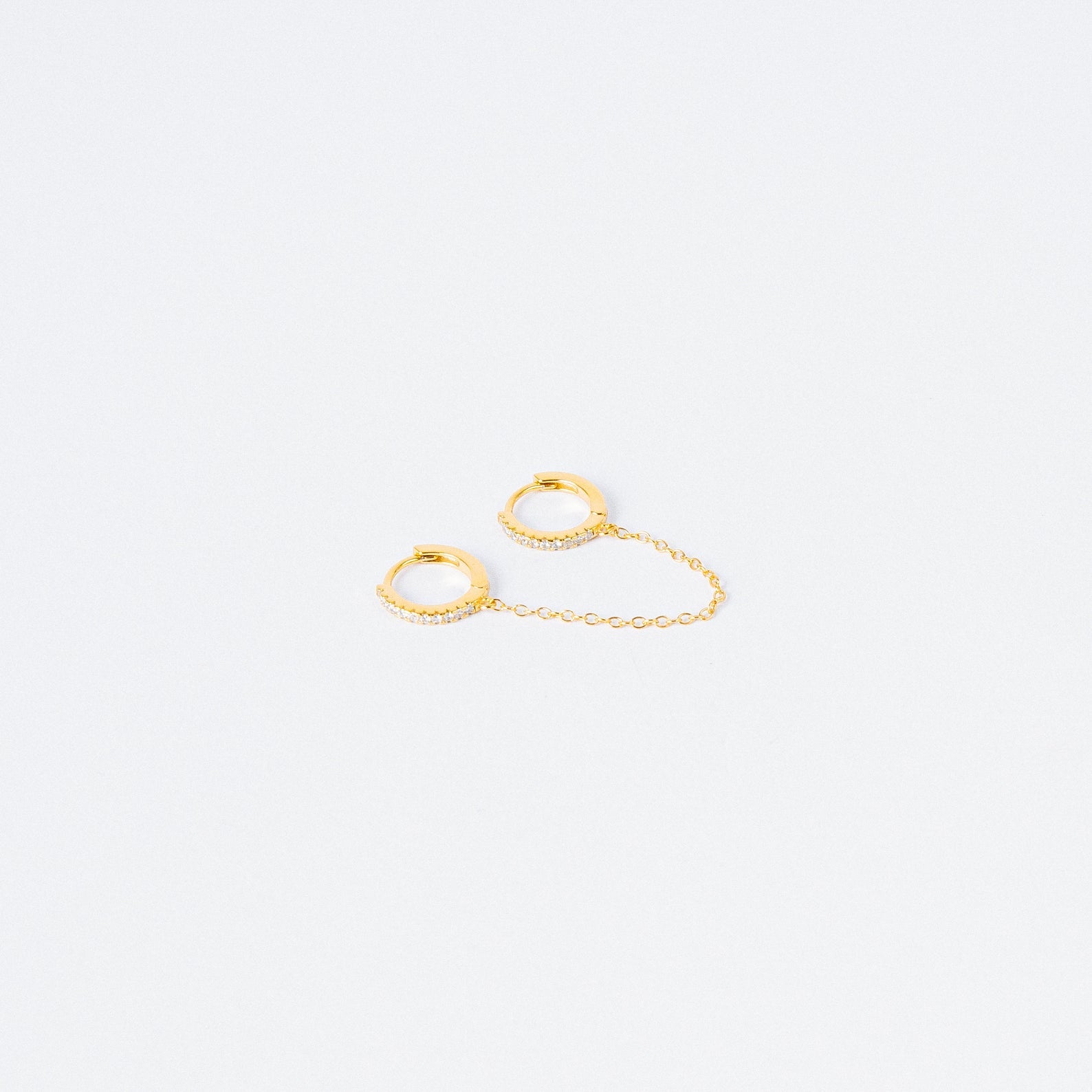The Pave Duo Chain Earrings feature two small gold huggies linked by a delicate chain, with one hoop embellished with clear stones—perfect for styling with multiple ear piercings.