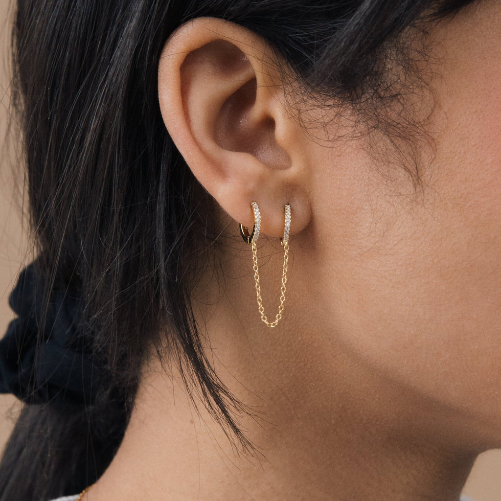 Close-up of a woman's ear with multiple piercings, showcasing double gold huggies linked by a fine chain, just like the Pave Duo Chain Earrings.