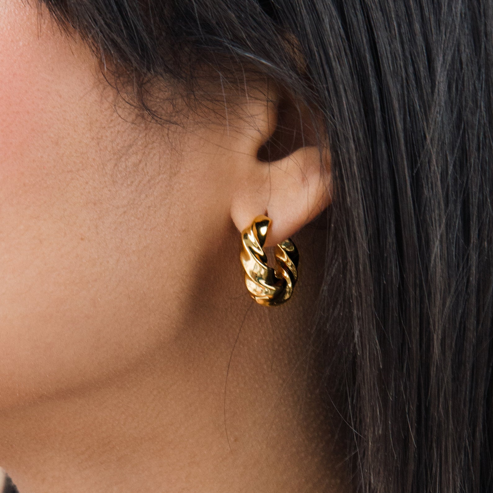 Close-up of a woman's ear with dark straight hair wearing the chunky gold Diana Hoops earring.