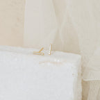 The Pave Bar Studs are minimal gold earrings with a sleek bar design and small pavé diamonds, displayed on a white textured block against a soft beige fabric background.
