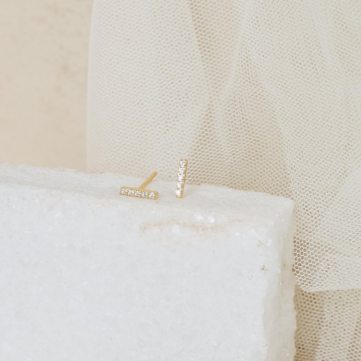 The Pave Bar Studs are minimal gold earrings with a sleek bar design and small pavé diamonds, displayed on a white textured block against a soft beige fabric background.
