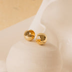 Chunky Ball Hoops, gold dome-shaped stud earrings inspired by the ‘70s, displayed on a smooth white ceramic surface with a soft background.