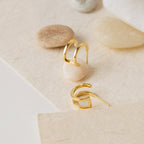 The Twin Hoops gold geometric earrings create a double earring illusion as they rest on a textured surface with smooth beige and tan stones.