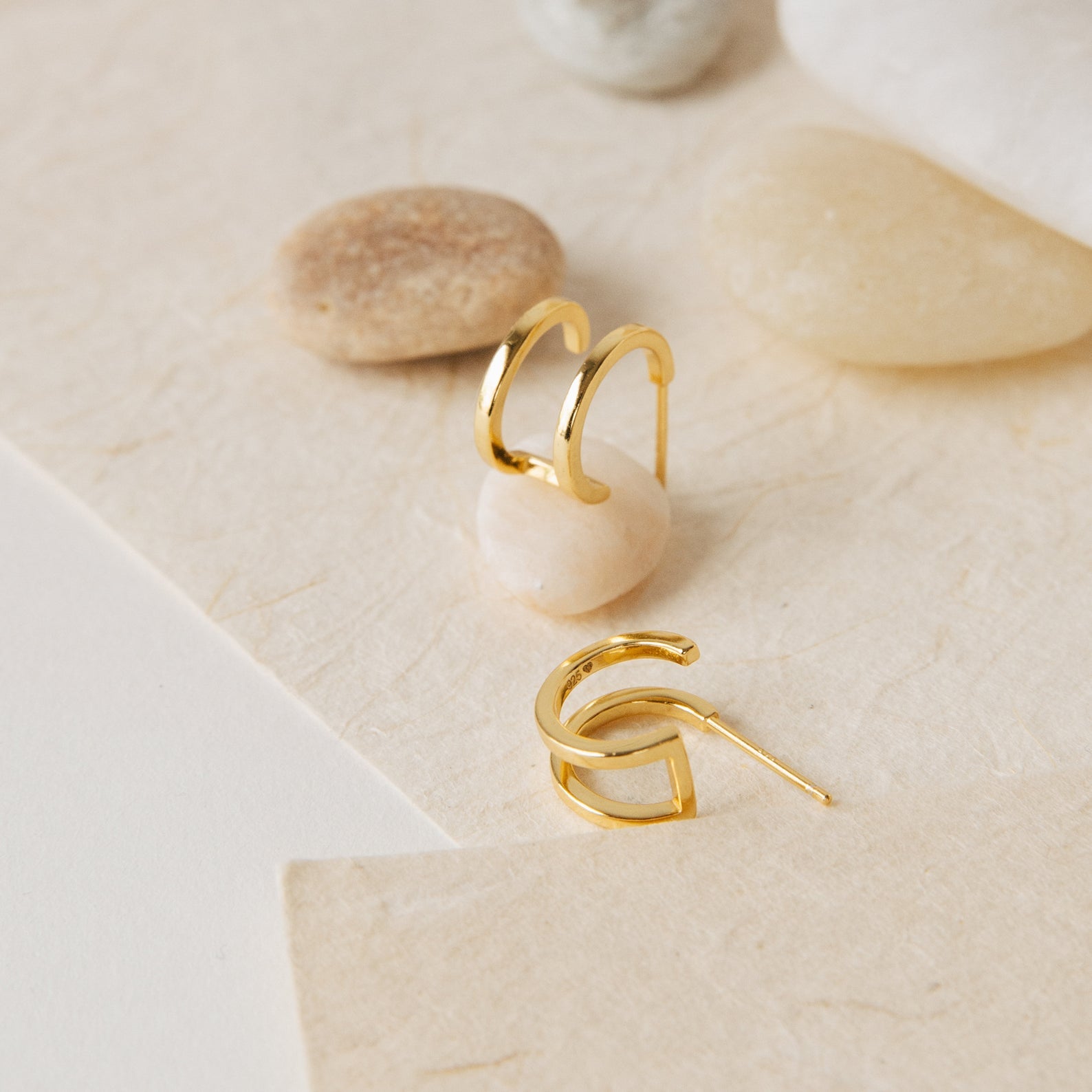 The Twin Hoops gold geometric earrings create a double earring illusion as they rest on a textured surface with smooth beige and tan stones.
