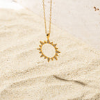 Gold sun-shaped pendant necklace resting on sand, featuring a hollow center and spiked edges embellished with pave set stones.