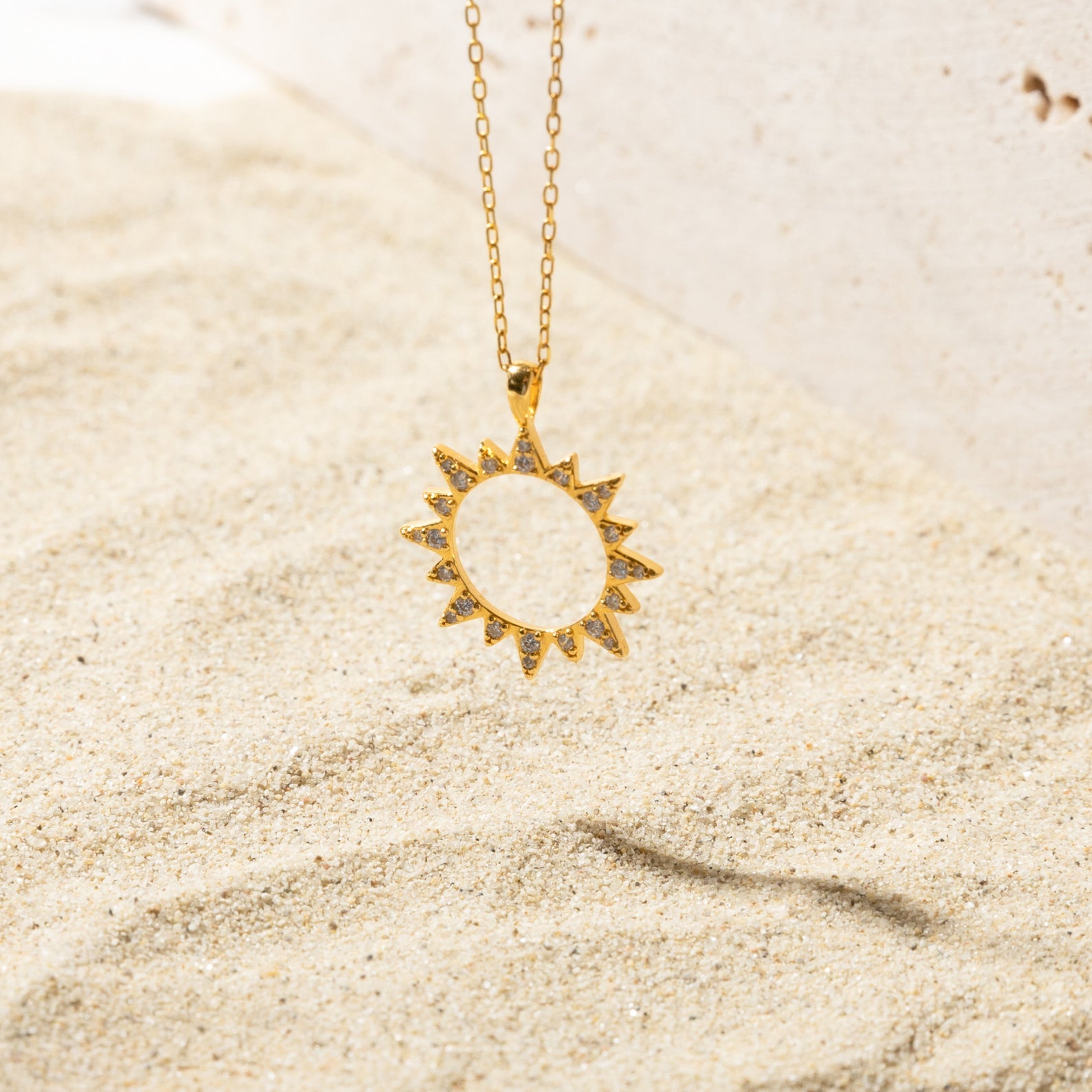 Gold sun-shaped pendant necklace resting on sand, featuring a hollow center and spiked edges embellished with pave set stones.